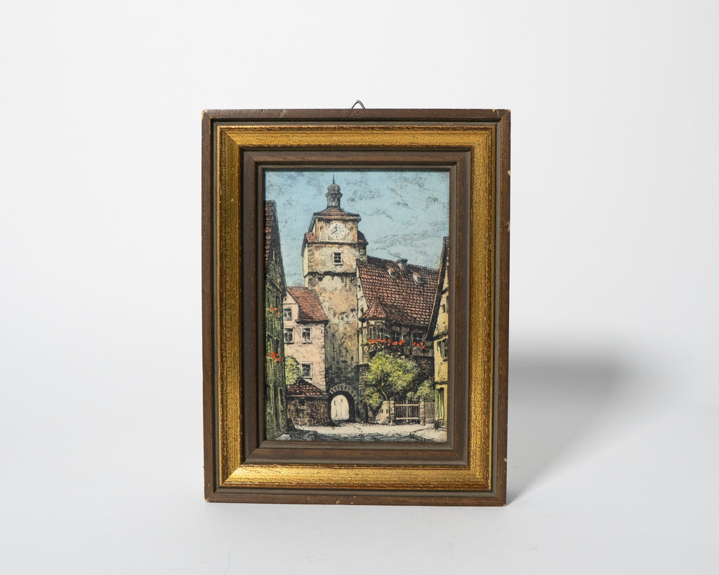 Framed painting of a village scene with a tower on a white background