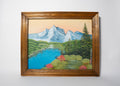 Framed painting of a mountain landscape with a lake and trees on a white wall.